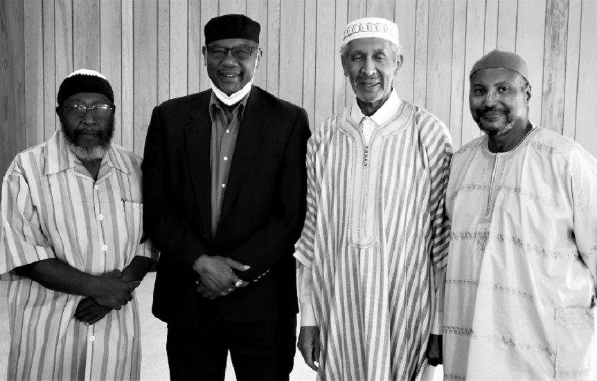 Four imams standing together in the wood-paneled musala, smiling, in kufis and robes.