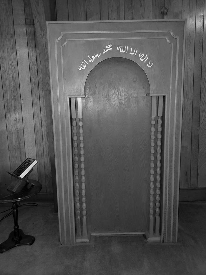 The wooden mihrab in the musala, framed by turned columns, with the shahada lettered above the arch.