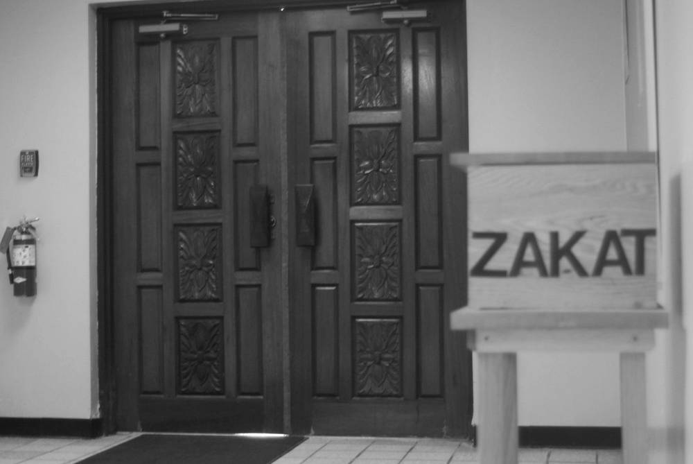 Carved wooden double doors inside the masjid, with a wooden box marked ZAKAT standing beside them.