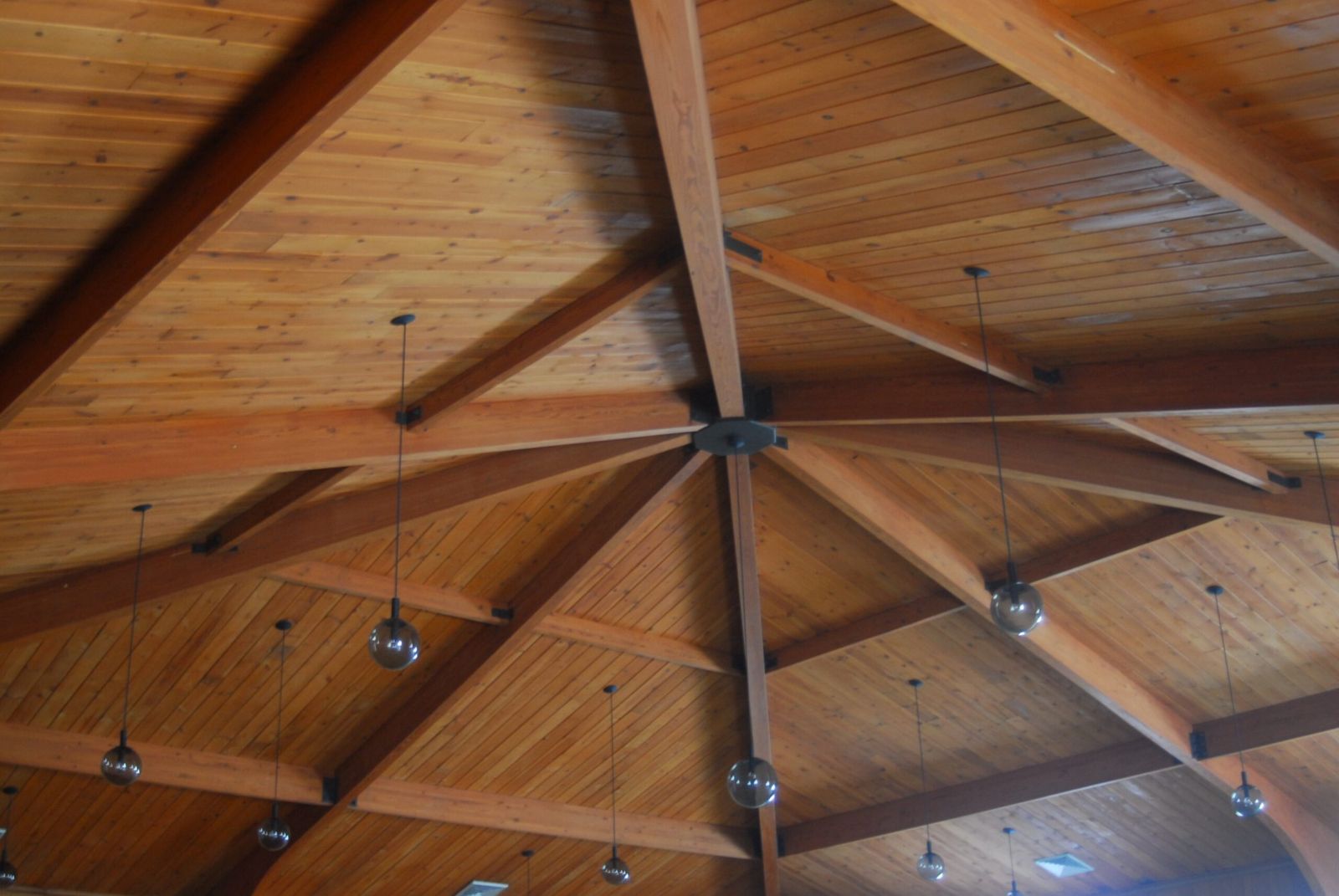 The timber roof beams of the musala, meeting at a central iron hub, with globe lamps hanging on long cords.