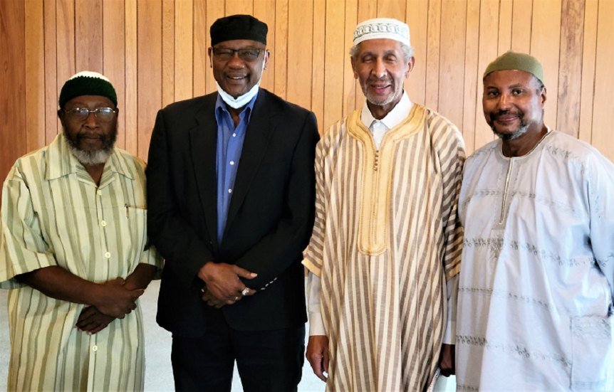 Four imams standing together in the wood-paneled musala, smiling, in kufis and robes.