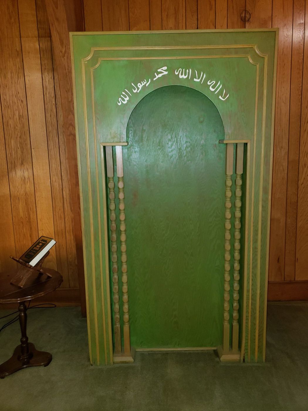 The wooden mihrab in the musala, framed by turned columns, with the shahada lettered above the arch.
