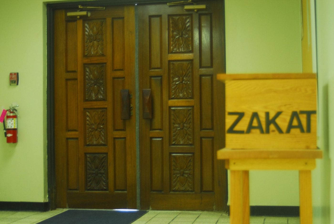 Carved wooden double doors inside the masjid, with a wooden box marked ZAKAT standing beside them.